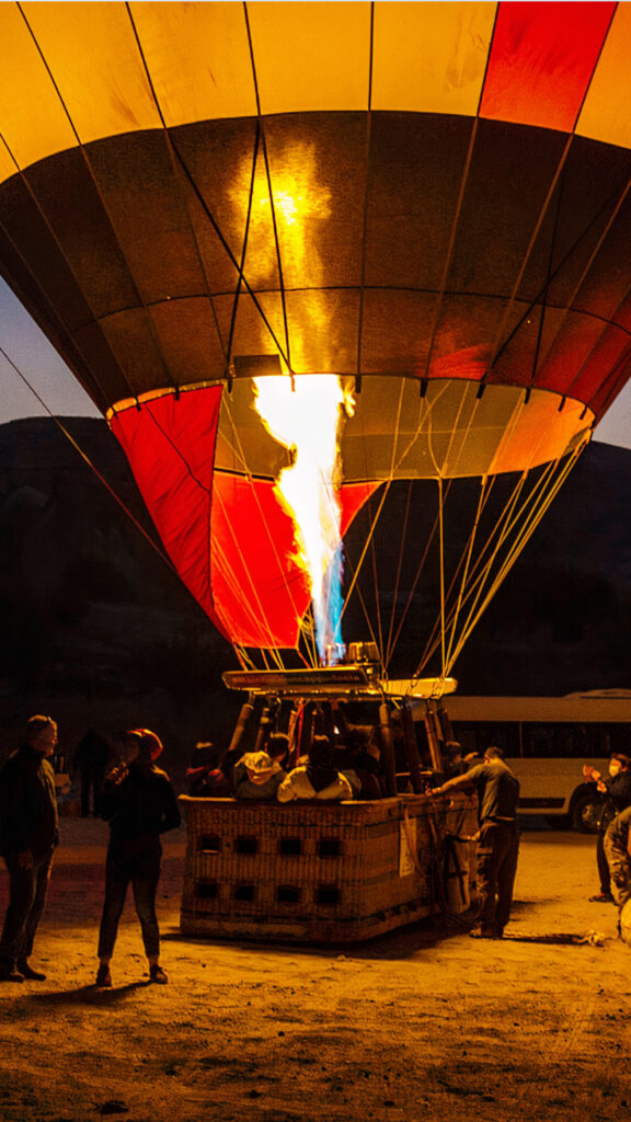 A hot air balloon being inflated at night, with a large flame emerging from the burner. The colorful balloon, with red, yellow, and black sections, is surrounded by a group of people preparing for the flight, creating a warm and dynamic scene under the darkening sky.