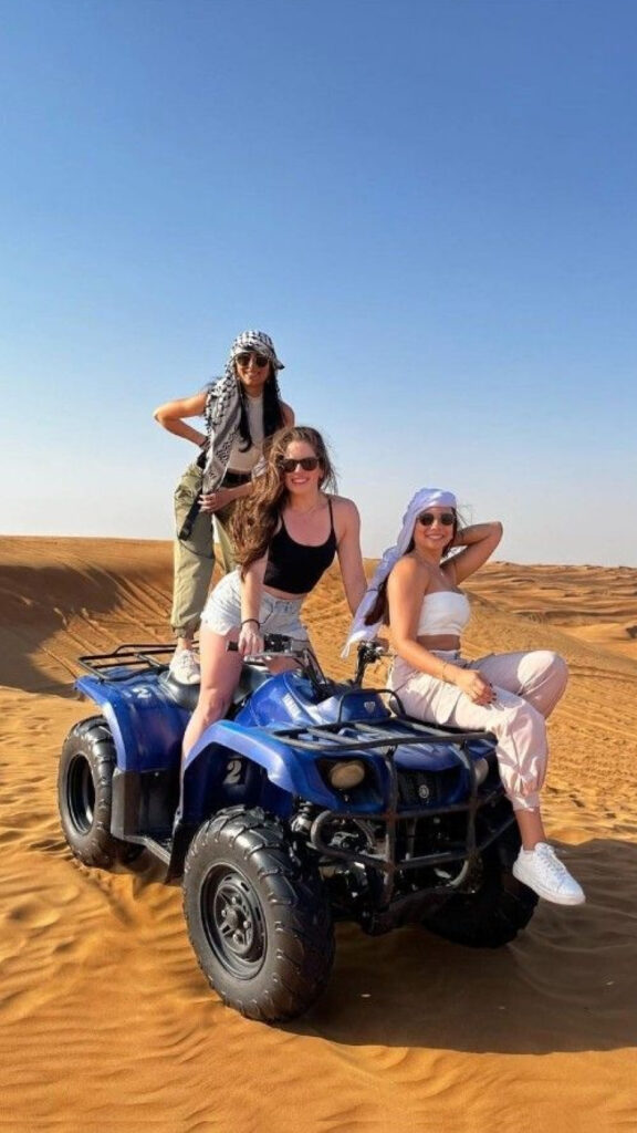Three women pose on a blue ATV in the desert. One stands on the vehicle, while the others sit, all wearing sunglasses and desert scarves, with the golden dunes and clear sky in the background.