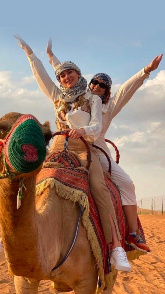 Two women joyfully riding a camel through the desert, both raising their arms in the air. They are wearing traditional headscarves and casual attire, with one woman in a white shirt and the other in a beige outfit, both smiling as they enjoy the ride.