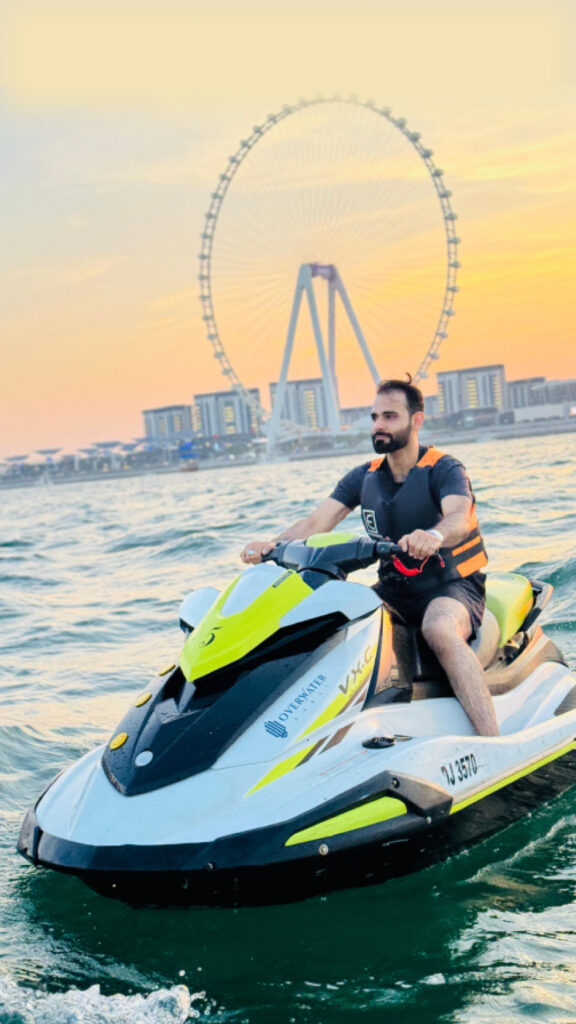 A person riding a jet ski in the water at sunset, with the Dubai Eye Ferris wheel visible in the background. The rider is wearing a life jacket and appears to be enjoying the calm waters, with a relaxed expression, as the golden hues of the sunset illuminate the scene.