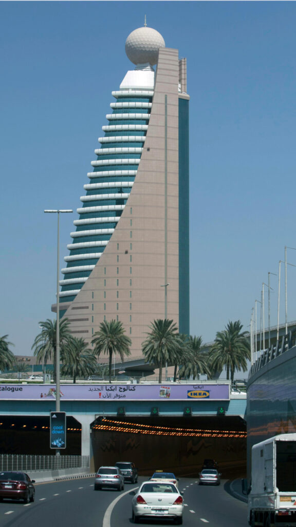 A distinctive modern tower with a round observation dome at the top, featuring a sleek, curved design and layered floors. The building stands tall against a clear blue sky, with palm trees lining the streets below, as cars drive through a tunnel under the structure.