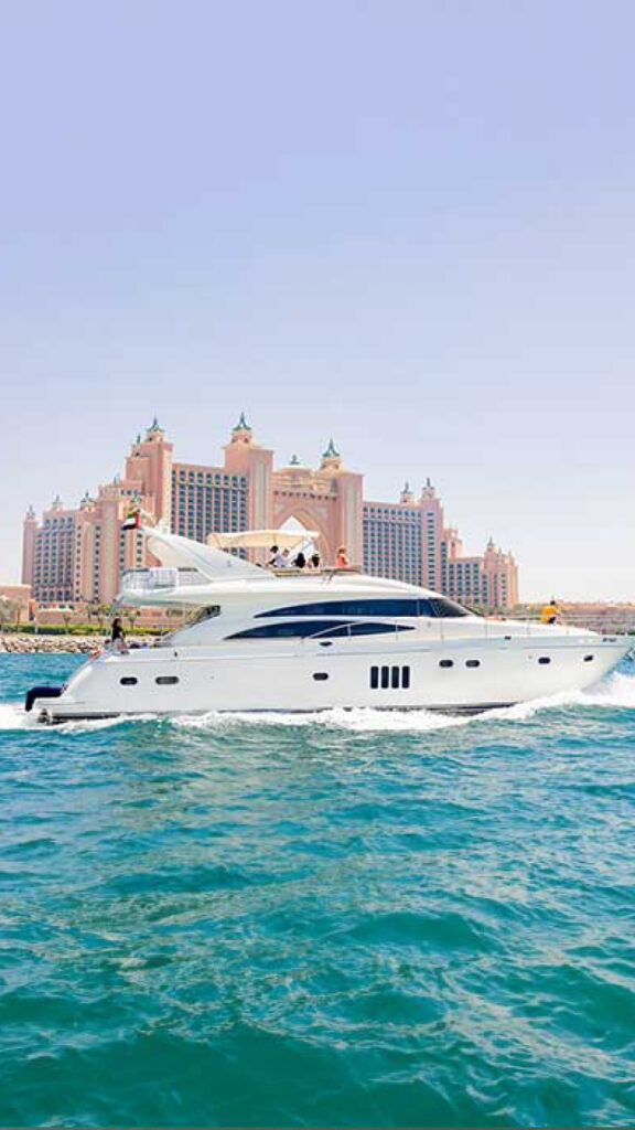 A luxurious white yacht cruising through clear blue waters, with several people visible on board. In the background, the iconic Atlantis hotel in Dubai stands tall, featuring its distinctive architecture, adding to the scene's grandeur."