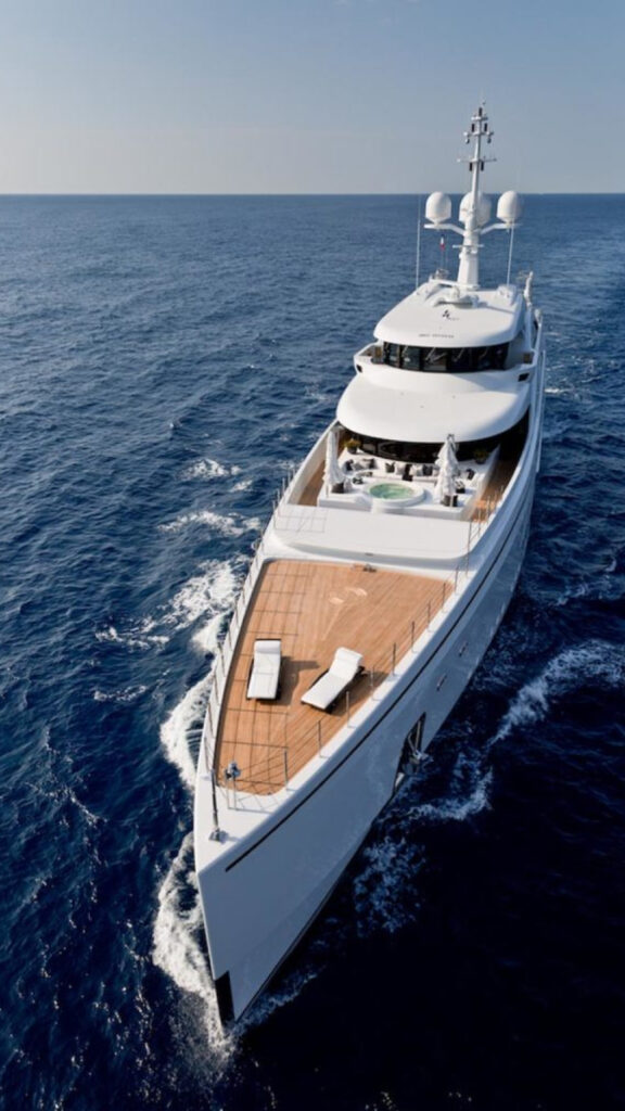 "A luxurious white yacht sailing on the open ocean with a large wooden deck. The yacht features modern design elements, including sunbeds and a round jacuzzi on the deck. The water is calm, and the sky is clear, emphasizing the yacht's elegance and exclusivity."