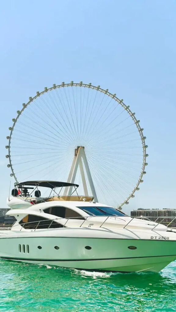 A sleek white yacht cruising on turquoise waters with the giant Ferris wheel, the Ain Dubai, towering in the background. The clear sky and modern architecture of the city add to the luxurious atmosphere of the scene."