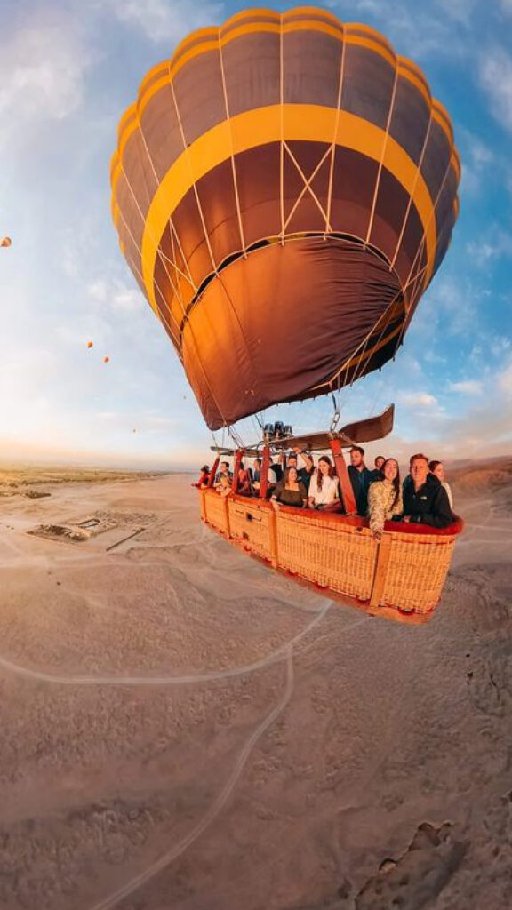 A vibrant hot air balloon soaring above the desert, with a group of people inside the basket, enjoying the aerial view. The balloon has a warm orange and yellow color scheme, and the expansive desert landscape stretches below, bathed in the soft light of dawn.