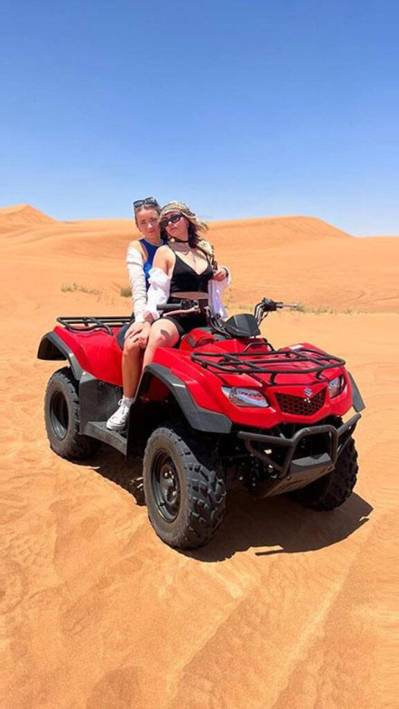 Two women pose on a red ATV in the desert, with one sitting at the front and the other standing behind. They both wear sunglasses and desert scarves, with golden sand dunes and a clear blue sky in the background.