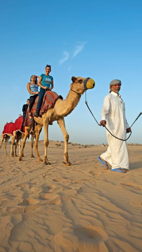 Two women riding camels through the desert, enjoying the view, while a man in traditional attire leads the camels. The scene is set under a clear blue sky, with the soft desert sand and distant dunes in the background.