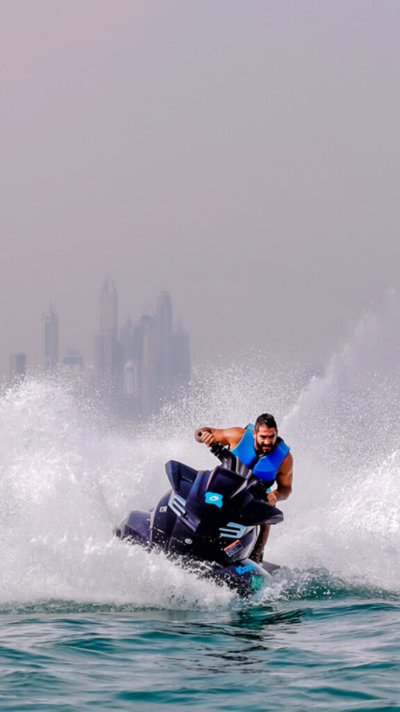A person riding a jet ski in the water, with tall buildings of Dubai's skyline visible in the background. The rider is wearing a blue life jacket and appears to be enjoying an adventurous ride, with water splashing around the jet ski.