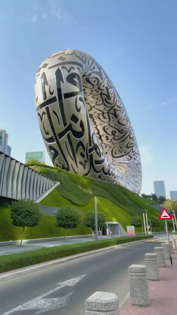 View of the Museum of the Future in Dubai, featuring its futuristic, ring-shaped architecture adorned with Arabic calligraphy. The building is surrounded by lush greenery, and the image captures the striking contrast against the clear blue sky.