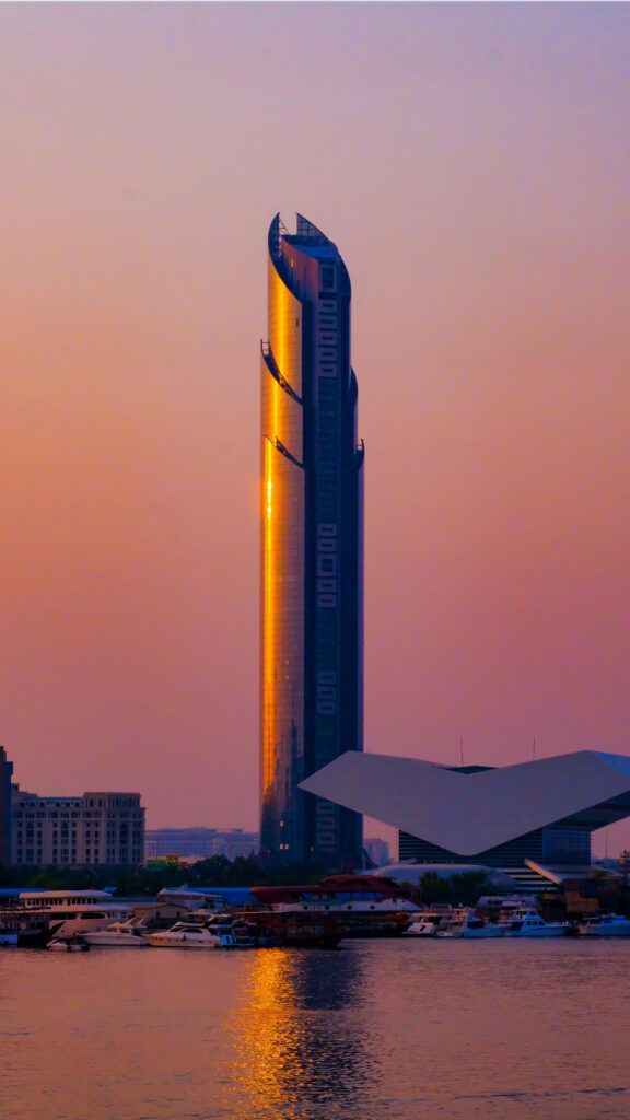 A modern skyscraper with a unique curved design, glowing under the warm colors of sunset. The building reflects golden light over the nearby water, with boats docked along the shore, creating a peaceful urban scene.