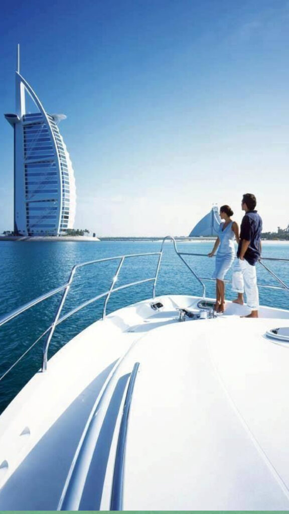 "A couple standing on the deck of a luxury yacht, looking out over the water towards the iconic Burj Al Arab hotel in Dubai. The sky is clear and blue, creating a serene atmosphere as the couple enjoys the scenic view."