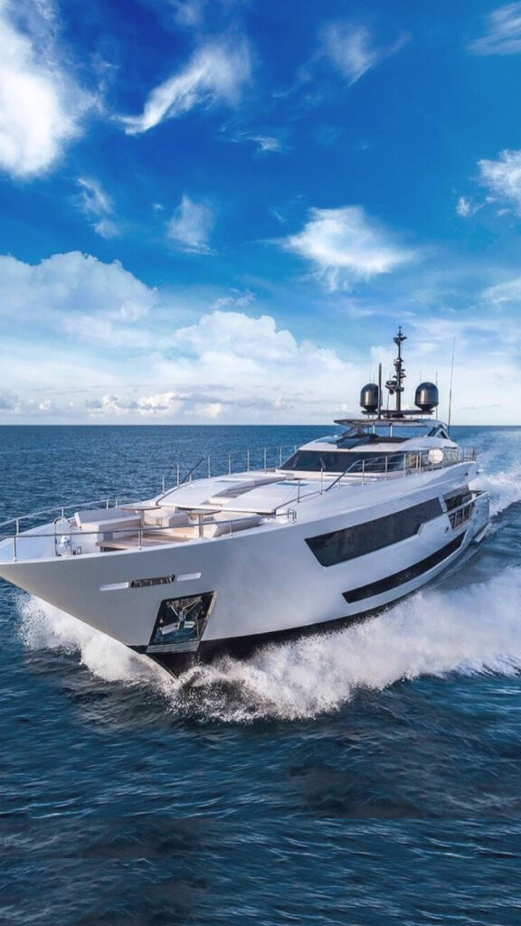 “A large modern luxury yacht speeding across open ocean, its white hull cutting through waves and throwing up spray, with a deep blue sea and a bright sky filled with scattered clouds.”