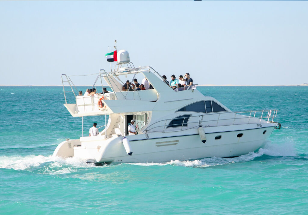 “A white motor yacht cruising on turquoise water with several passengers gathered on the upper deck, a crew member visible at the rear, and a national flag flying above, with a clear sky and distant shoreline in the background.”