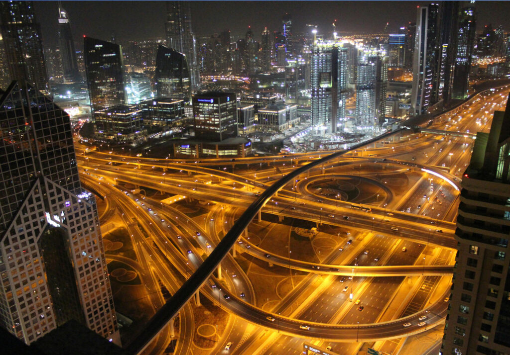 A bird’s eye view of a vibrant, illuminated cityscape at night. The bustling highway is filled with moving cars, and the intricate highway interchange curves in glowing yellow lights. Tall skyscrapers and office buildings light up the skyline, with construction sites in the distance, adding to the dynamic energy of the scene. The lights from the roads and buildings create a lively contrast against the dark sky.