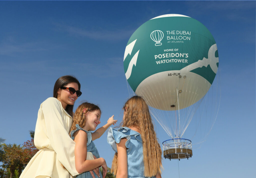 A woman and two children stand below the Dubai Balloon at Atlantis, looking up with excitement. The vibrant green balloon with the logo 'The Dubai Balloon' and the tagline 'Home of Poseidon's Watchtower' is floating high in the clear blue sky.