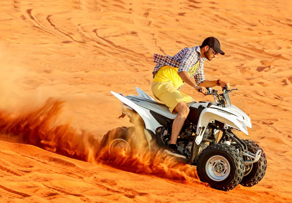A man riding a white ATV through the desert, kicking up a cloud of sand as he speeds across the dunes. He wears a yellow shirt, shorts, a plaid shirt, and a cap, with sunglasses for protection from the sun.