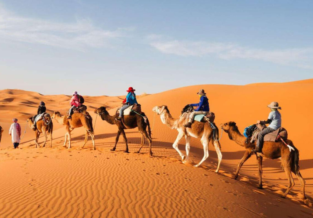 A group of tourists riding camels across the vast orange sand dunes of the desert. The guide, wearing traditional attire, leads the camels while the riders, dressed in colorful jackets and hats, enjoy the scenic desert landscape.