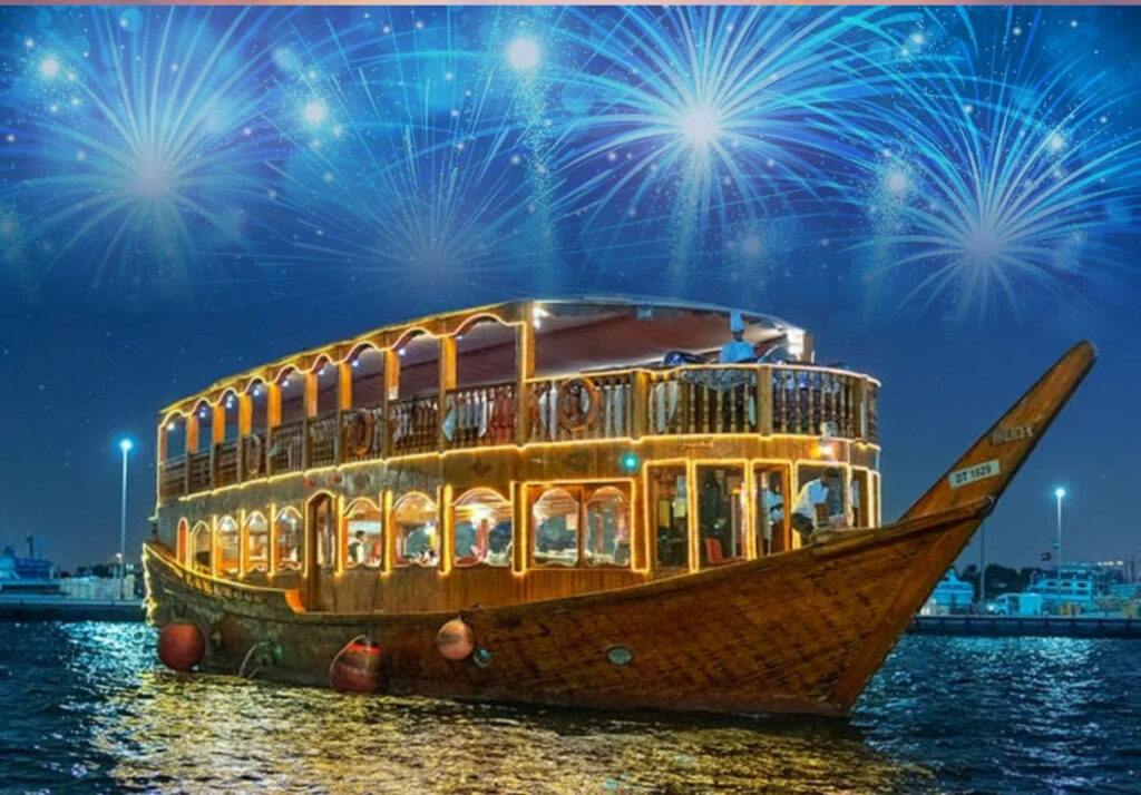 A traditional wooden dhow boat lit with vibrant lights, sailing on calm waters at night. The boat is decorated with festive lights and has people on board, some visible through the windows. The background features a sky filled with colorful fireworks, adding to the celebratory atmosphere