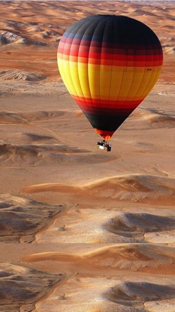 A vibrant hot air balloon with stripes of red, yellow, and black soaring above the vast desert landscape. The balloon hovers gently over the rolling dunes, casting a striking contrast against the warm, golden sands of the desert below.