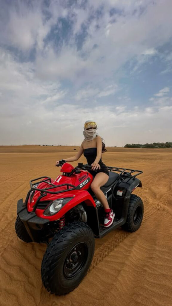 A woman wearing a scarf and sunglasses rides a red ATV across the desert sand dunes. The ATV is labeled '250' on the side, and the woman is dressed in a black outfit with red sneakers. The sky above is partly cloudy, and the desert landscape stretches into the distance.