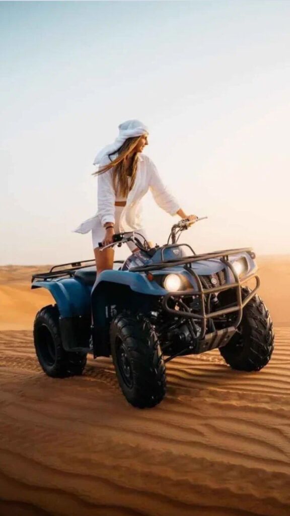 "A woman in a white outfit and headscarf riding an ATV across the desert sands, with her hair flowing in the wind and the vast desert landscape stretching out behind her under a clear sky.