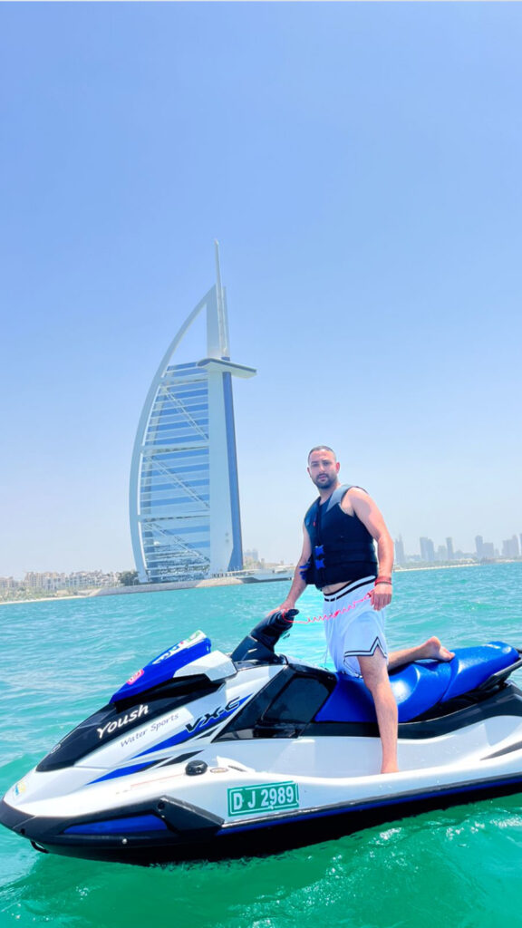 A person standing on a jet ski in the clear waters of Dubai, with the Burj Al Arab hotel in the background. The rider is wearing a life jacket and casual swimwear, looking relaxed and confident as they enjoy the iconic view of the city’s skyline.