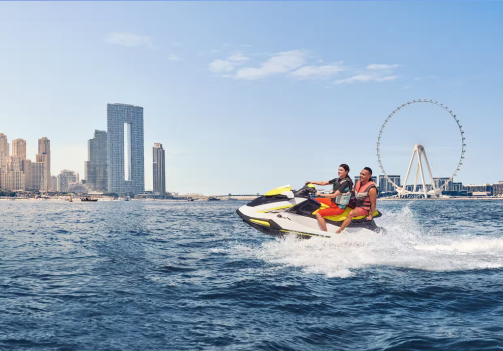 Two people riding a jet ski together in the waters near Dubai, with the Dubai Eye Ferris wheel visible in the background. The riders are wearing life jackets and appear to be enjoying the adventure, surrounded by tall buildings on the coastline under a bright, clear sky.