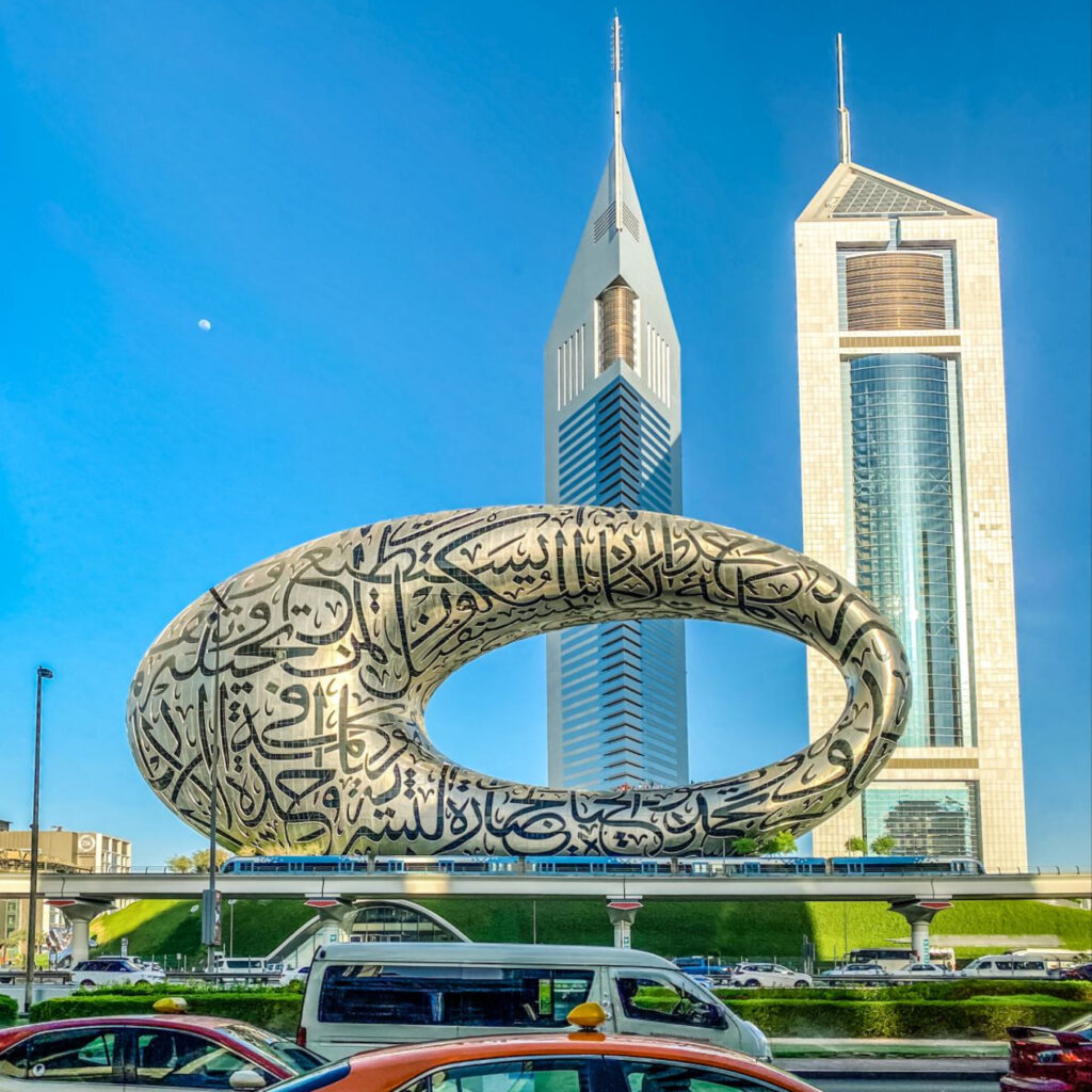 The image showcases the Museum of the Future, an innovative and striking building in Dubai. The museum is a large, futuristic oval structure covered in Arabic calligraphy, set against a bright blue sky. In the background, two tall, modern skyscrapers rise, adding to the city's contemporary skyline. The scene captures the intersection of advanced architecture with urban life, with cars and taxis visible in the foreground.